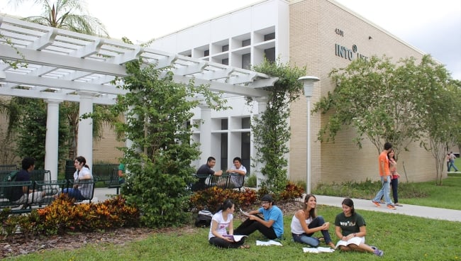 A group of students sits on a lawn outside a cream-colored building